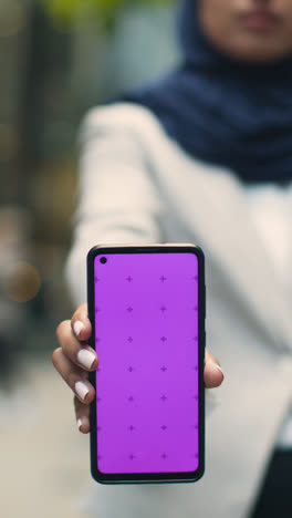 Vertical-Video-Close-Up-Of-Muslim-Businesswoman-Holding-Mobile-Phone-With-Blank-Screen-To-Camera-Standing-Outside-Office-In-City